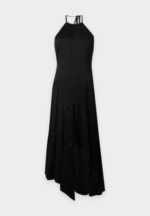 Femme portant une robe noire sans manches à col halter et des sandales noires à talons hauts, debout sur un fond blanc.