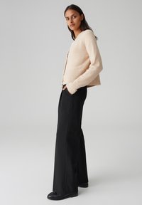 Cardigan en laine beige clair avec un ourlet côtelé, fermeture à boutons, porté sur un pantalon large noir. Texture douce, coupe décontractée.