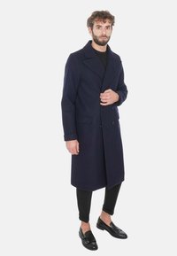 Cappotto di lana blu navy con ampio colletto, bottoni doppi e tasche laterali, abbinato a pantaloni e scarpe neri.