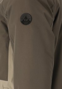 Grüne Outdoor-Jacke mit beigem Seitenpanel, ausgestattet mit einem runden schwarzen Patch mit Bergdesign und dem Markennamen "Whistler". Glatte Stofftextur.