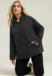 Veste noire imperméable avec capuche, dotée d'une fermeture à bouton, de deux poches à l'avant et de poignets côtelés, portée sur un haut blanc et un pantalon noir.