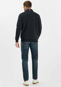 Uomo in piedi di spalle, indossa una maglietta nera a maniche lunghe, jeans blu e sneakers bianche, su uno sfondo bianco semplice.