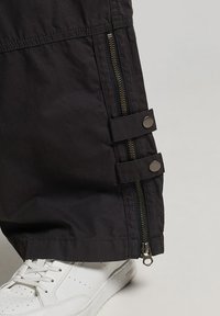 Zwarte oversized broek met ritsen en drukknoopsluitingen aan de pijpen, gemaakt van een gladde, lichte stof, gecombineerd met witte sneakers.