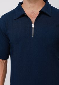 Marineblaue Kurzarm-Polohemd mit strukturiertem Rautenmuster und silbernem Reißverschluss am Hals. Verfügt über einen Spitzenkragen und subtile Stickdetails.