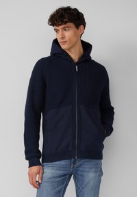 Navy Kapuzenzipperpullover mit geripptem Stoff, zwei vorderen Taschen und kontrastierendem Stoff am unteren Vorderteil, kombiniert mit hellblauen Jeans.