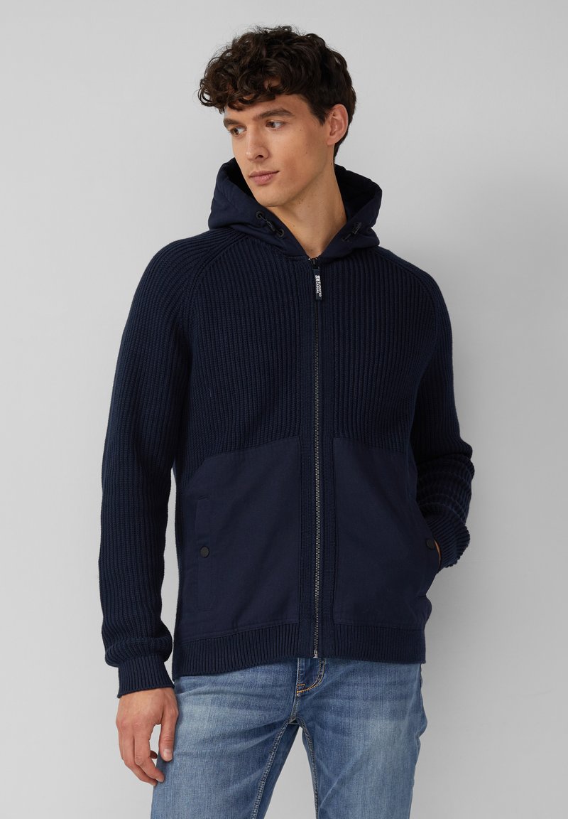 Navy Kapuzenzipperpullover mit geripptem Stoff, zwei vorderen Taschen und kontrastierendem Stoff am unteren Vorderteil, kombiniert mit hellblauen Jeans.