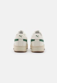 Une paire de baskets Puma blanches avec des rayures et des logos verts, présentée de dos sur un fond blanc uni.