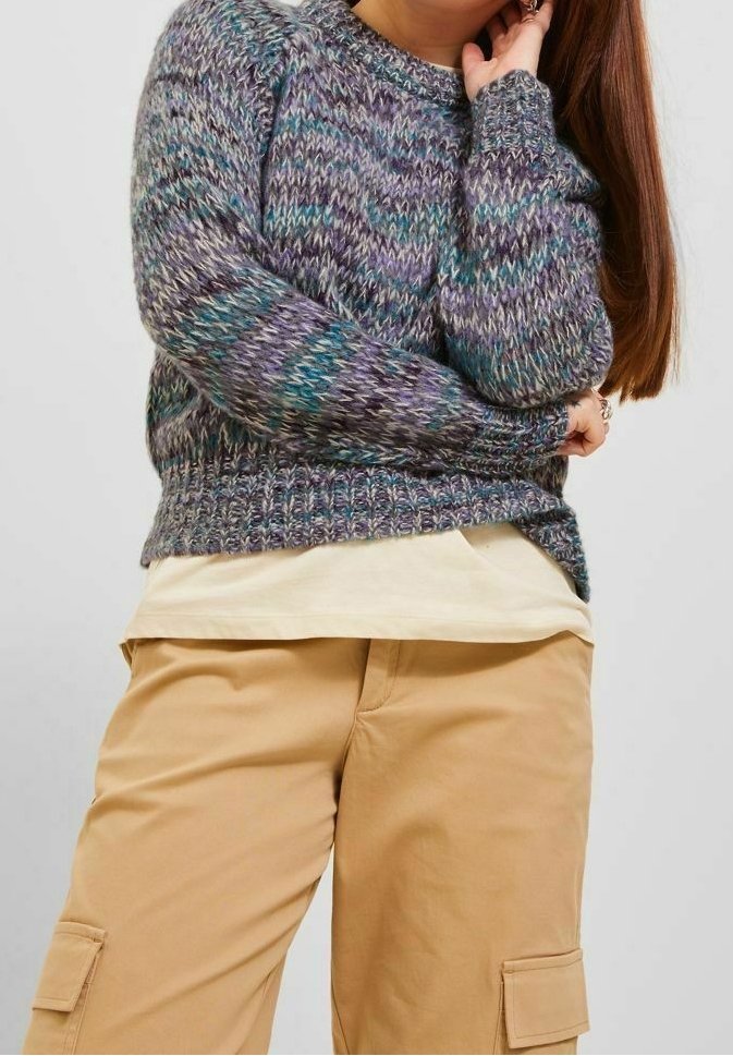 Personne portant un pull en maille violet et sarcelle par-dessus une chemise beige et un pantalon cargo beige, posant avec une main près du visage.