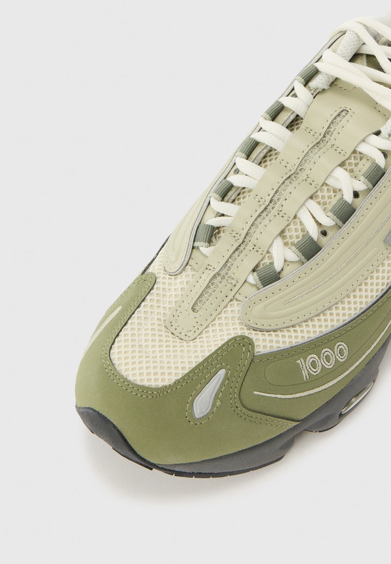 Zapatilla de color verde oliva con paneles de malla, acentos texturizados y suela gruesa. Presenta cordones blancos y un detalle de "1000" en un color contrastante.
