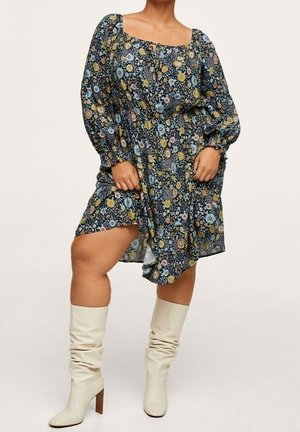 Mujer con un vestido azul marino con estampado floral, mangas largas abullonadas y botas crema hasta la rodilla, de pie con una pierna ligeramente levantada.