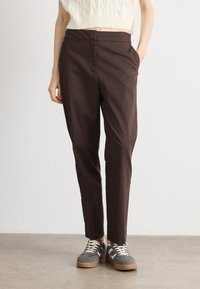 Brown tapered trousers with side pockets, made of smooth fabric. Paired with grey and beige sneakers featuring a textured design.