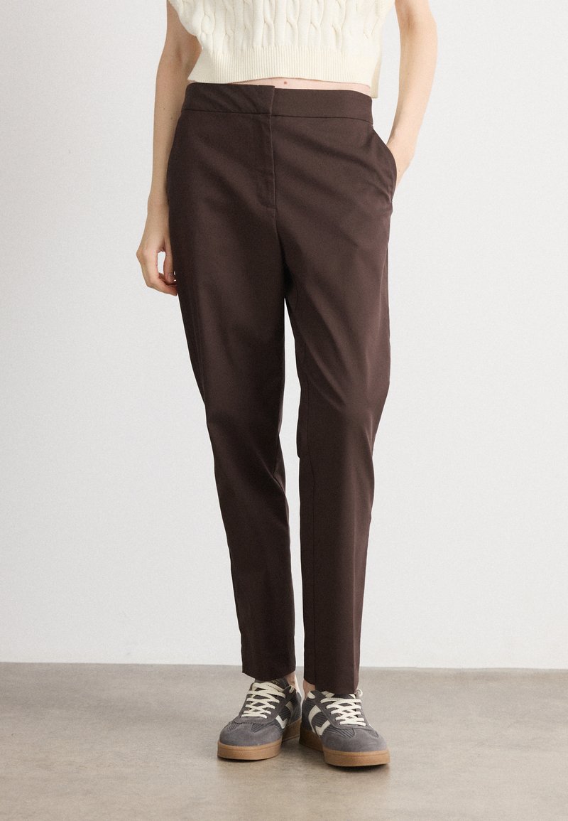 Brown tapered trousers with side pockets, made of smooth fabric. Paired with grey and beige sneakers featuring a textured design.