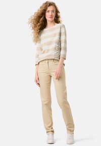 Beige striped sweater with three-quarter sleeves and text detail, paired with beige pants and white sneakers. Model stands against a plain background.