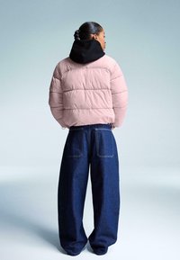Giacca a vento rosa chiaro con motivo a coste orizzontali, cappuccio nero, abbinata a jeans di denim blu scuro a gamba larga. Tessuto liscio e vestibilità rilassata.