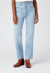 Persona vestida con jeans rasgados de color azul claro y botas de cuero marrón hasta el tobillo, de pie contra un fondo blanco liso.