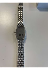 Srebrni metalni sat s pravokutnim kućištem, povezanom narukvicom i ravnim metalnim stražnjim dijelom s ugraviranim markama branda i certifikacije.