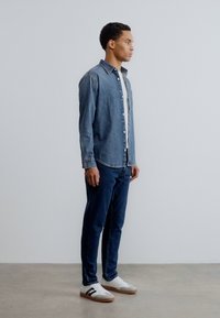 Πουκάμισο από τζιν σε σκουρό μπλε χρώμα, με κούμπωμα κουμπιών· συνδυασμένο με μπλε τζιν και λευκά αθλητικά παπούτσια με μαύρες λεπτομέρειες. Απλή, μοντέρνα εφαρμογή.