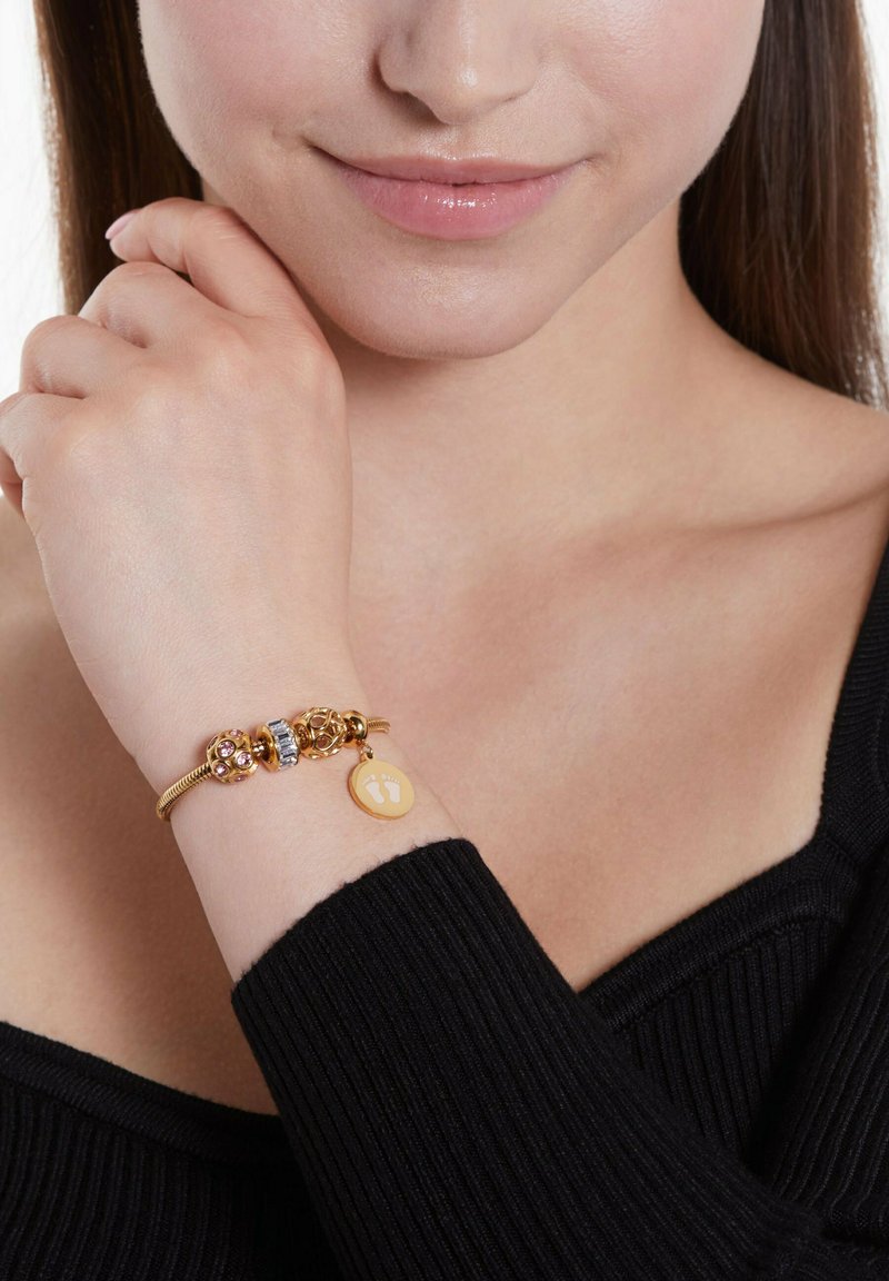 Goldener Armband mit dekorativen Charms und rosa Steinen sowie einem runden gelben Tag mit Fußabdrücken. Am nackten Handgelenk getragen.