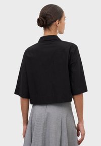 Mujer con el cabello recogido en un moño, que lleva una chaqueta negra corta con mangas hasta el codo y una falda plisada gris texturizada, de espaldas contra un fondo liso.