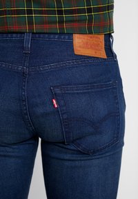 Jeans de mezclilla de color azul oscuro presentan un parche de cuero con el logo de la marca, detalles en el bolsillo trasero y acentos de costura dorada. Se llevan debajo de una camisa de cuadros.