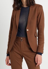 Brun kavaj med en frontficka, guldknappdetaljer och skräddarsydd passform, i kombination med en mörkblå topp med prickar och matchande byxor.