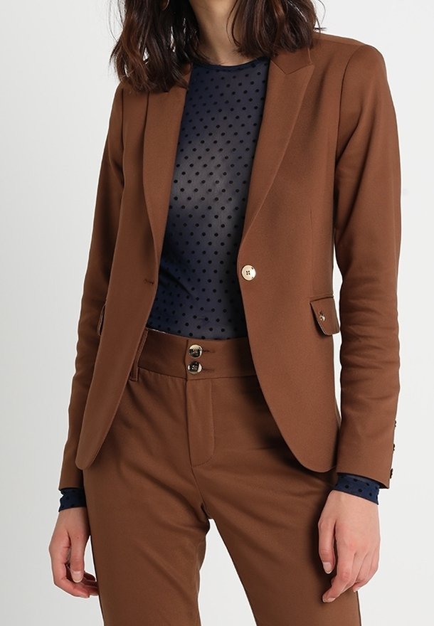 Brun kavaj med en frontficka, guldknappdetaljer och skräddarsydd passform, i kombination med en mörkblå topp med prickar och matchande byxor.