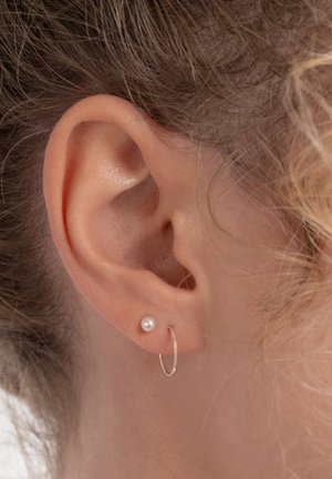Gouden oorring in hoepelvorm en een kleine parelsteek aan het oor van een persoon, met een gladde textuur en een eenvoudig ontwerp tegen een lichte huid en haar.