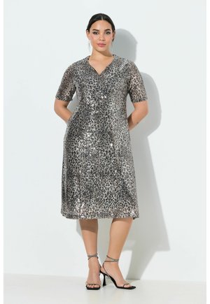 Kvinde stående op ad en hvid væg iført en knælængde, kortærmet sølvglitrende palietkjole og sorte højhælede sandaler med stropper.