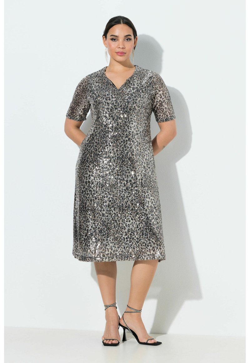 Žena stojící opřená o bílou zeď, oblečená v stříbrných flitrových šatech s délkou ke kolenům a krátkými rukávy, a v černých lodičkách s pásky.