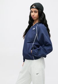 Γαλάζιο αντιανεμικό μπουφάν με λευκές ραφές, ελαστικές μανσέτες και τελείωμα· διαθέτει το λογότυπο της Nike στο στήθος. Συνδυάζεται με ανοιχτό γκρι φόρμα παντελόνι.