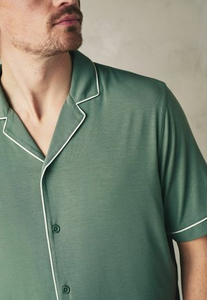 Mann trägt grünes, kurzärmeliges Hemd mit weißen Ziernähten, sichtbar vom Hals bis zur Mitte des Oberkörpers vor neutralem Hintergrund.