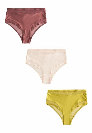 Vrouw met schouderlang blond haar, gekleed in een geelgroene doorschijnende mesh beha en hooggetailleerde slip, staand tegen een effen lichte muur.