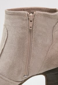 Bottine en suède beige avec une fermeture éclair sur le côté, finition texturée et talon empilé, présentant des détails de couture et un bout arrondi.