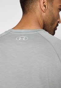 Γκρι αθλητικό μπλουζάκι με υφή υφάσματος, στρογγυλή λαιμόκοψη και μανίκια ράγκλαν. Το λογότυ πο της Under Armour είναι ορατό στο πίσω μέρος του λαιμού.