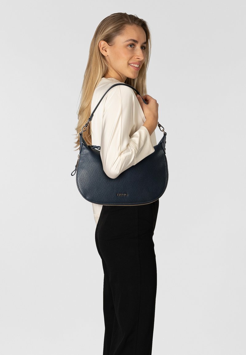 Borsa a tracolla in pelle blu navy con finitura texturizzata, caratterizzata da una forma arrotondata e una tracolla con dettagli a catena. Dettaglio logo dorato sulla parte anteriore.