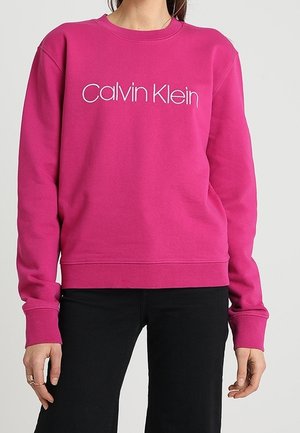 Osoba nosząca jasno różową bluzę Calvin Klein i czarne spodnie, z rękami swobodnie opadającymi wzdłuż ciała.