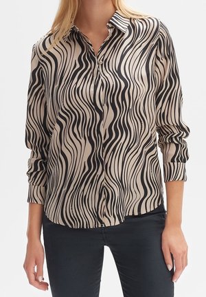 Frau trägt eine beige langärmelige Bluse mit schwarzen wellenförmigen Streifen und schwarze Hose.