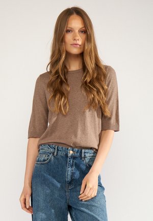 Jeune femme aux longs cheveux ondulés porte un pull marron à manches courtes et un jean bleu taille haute, debout devant un fond uni.