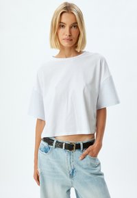 Weißes, kurzes T-Shirt mit blauen gestreiften kurzen Ärmeln, rundem Ausschnitt und weichem Stoff. Getragen mit hellblauen, abgewetzten Jeans und einem schwarzen Gürtel.