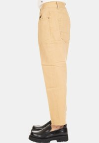 Pantaloni beige a sigaretta indossati con mocassini neri con zeppa, mostrati in vista laterale su uno sfondo chiaro e semplice.
