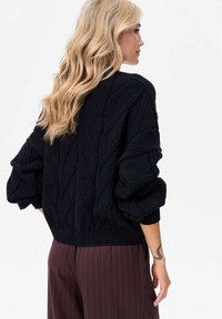 Czarny sweter z dzianiny z teksturowanym wzorem diamentowym i luźnymi, objętościowymi rękawami, w zestawieniu z brązowymi spodniami w paski.