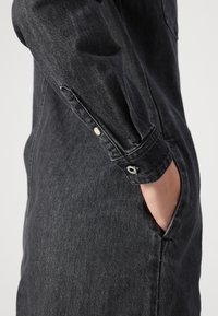 Čierne deníny šaty s golierom, dlhými rukávmi so zapínaním na cvoky a bočnými vreckami. Štruktúra je mierne textúrovaná a odolná.