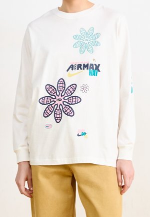 Biała koszula z długim rękawem z kolorowymi kwiatowymi wzorami i logo Nike Air Max, noszona z beżowymi spodniami, pokazana od ramion do bioder.