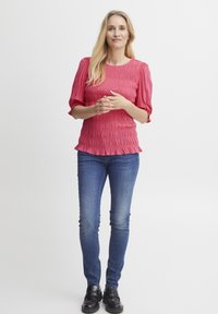 Rozā smocked tops ar īsām, pūkainām piedurknēm, kas apvienots ar zilām plānām biksēm un melniem apaviem. Vienkāršs, teksturēts dizains ar ruķinājuma detaļām.