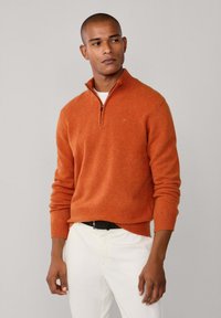 Maglione arancione con zip e collo alto in un morbido knit, maniche lunghe, con polsini e vita a costine, abbinato a pantaloni bianchi e una cintura nera.