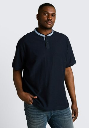 Mann mit kurzen Haaren trägt ein marineblaues Henley-Shirt mit kurzen Ärmeln und hellblauem Kragen, steht mit einer Hand in der Tasche vor einfarbigem Hintergrund.