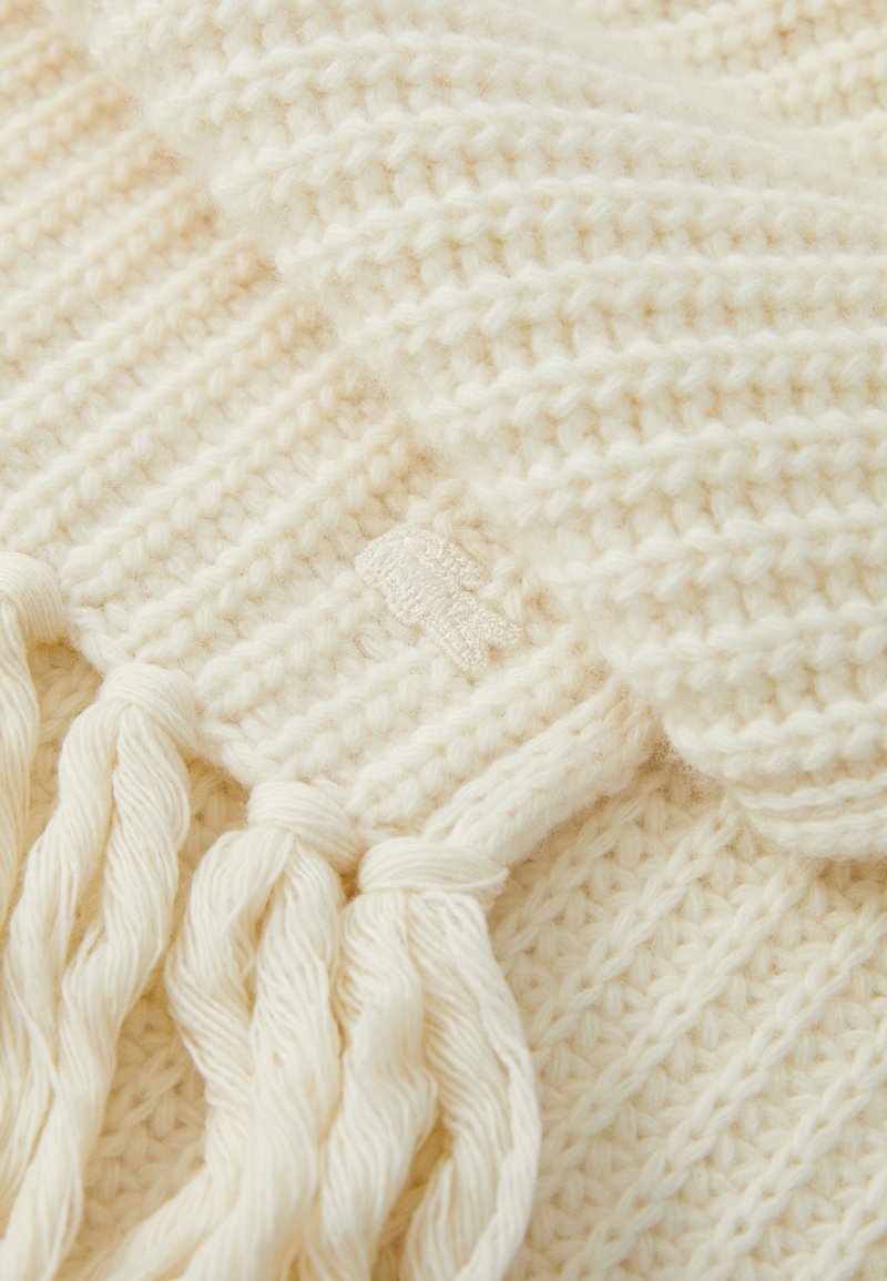 Gestrickter elfenbeinfarbener Schal mit einem strukturierten Muster, ausgestattet mit verdrehten Fransen an den Enden und einem kleinen, gestickten Etikett am Rand. Weiches, warmes Material.