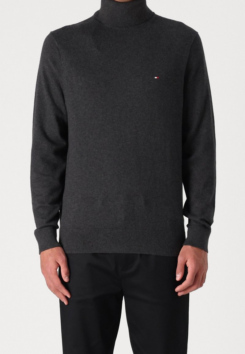 Homme portant un pull à col roulé gris foncé avec un petit logo sur la poitrine et un pantalon noir, debout devant un fond uni.