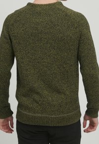 Man met een groen en zwart gemarmerd gebreid trui gezien vanuit de rug, gecombineerd met zwarte broek tegen een effen achtergrond.