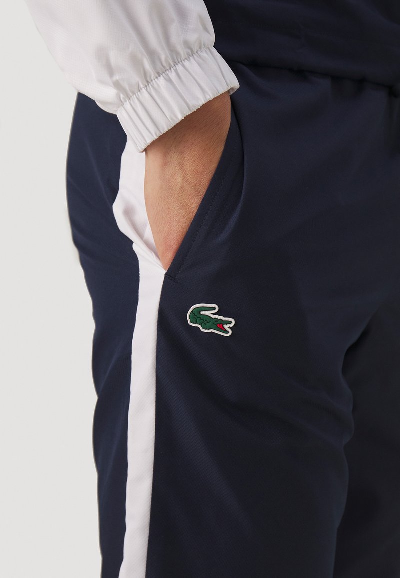Pantalones joggers en color navy con cintura elástica, bolsillos laterales y franjas blancas en los costados. Presenta un logo de cocodrilo verde de Lacoste en el muslo.